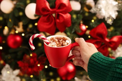 Woman holding cup of delicious Christmas cocoa with marshmallows and candy cane near decorated fir tree, closeup