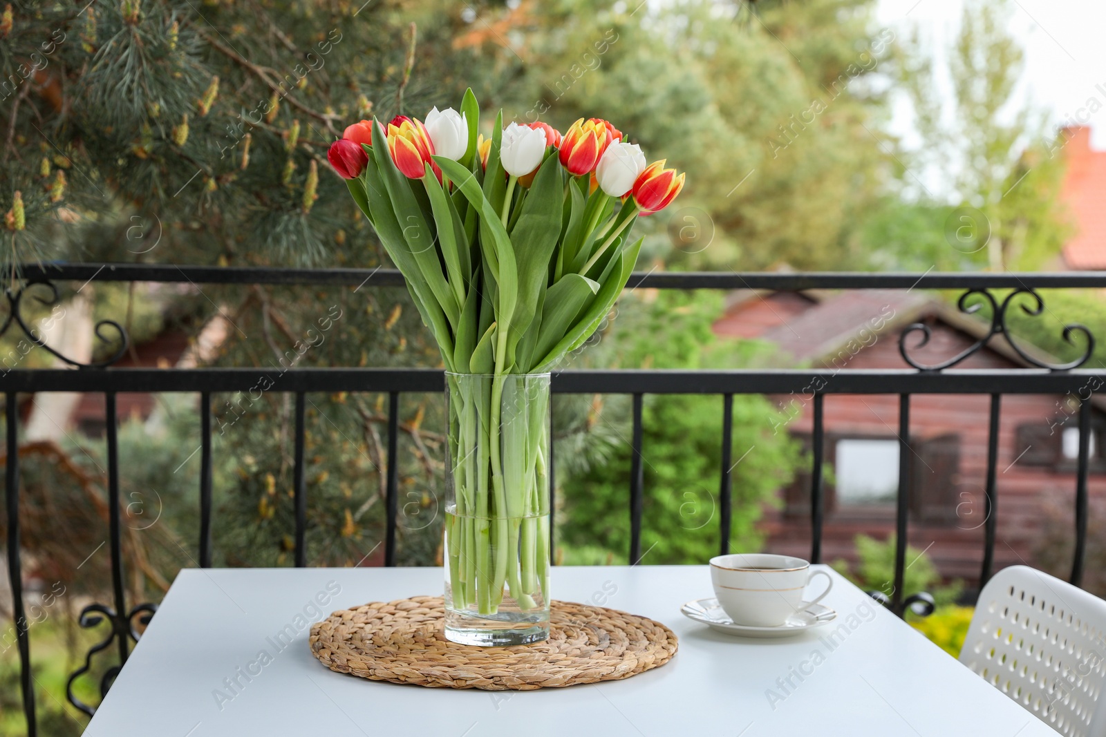 Photo of Beautiful colorful tulips in glass vase and cup of drink on white table at balcony