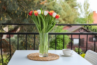 Beautiful colorful tulips in glass vase and cup of drink on white table at balcony