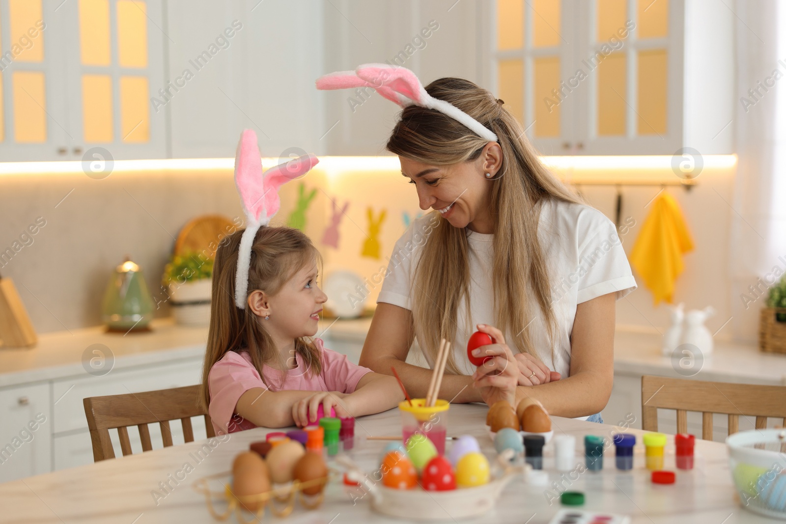 Photo of Easter celebration. Mother with her cute daughter painting eggs at white marble table in kitchen