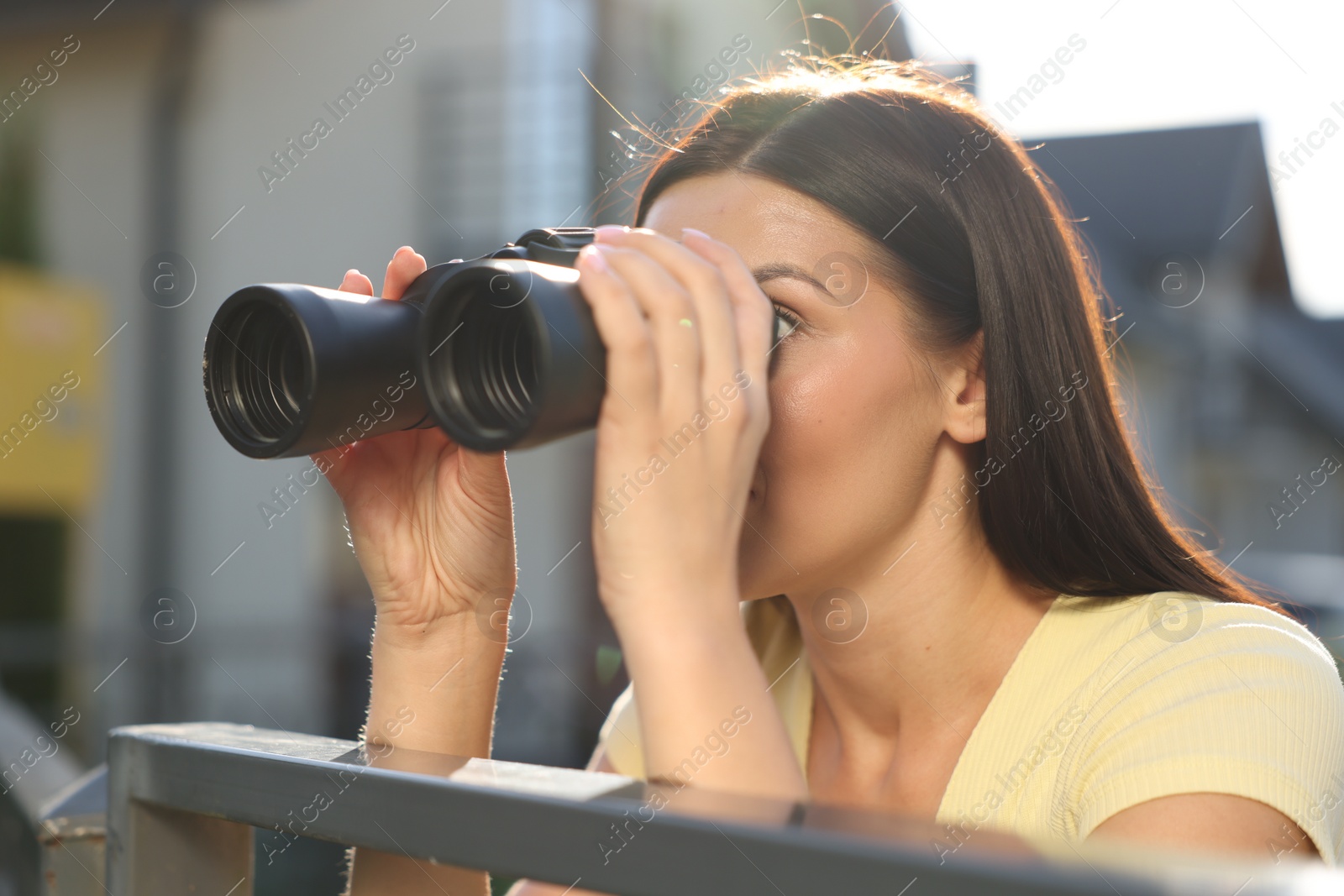 Photo of Concept of private life. Curious young woman with binoculars spying on neighbours over fence outdoors