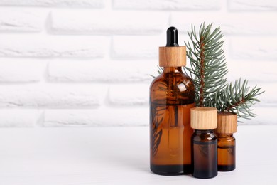 Photo of Bottles of essential oil and pine branch on white wooden table near brick wall, space for text