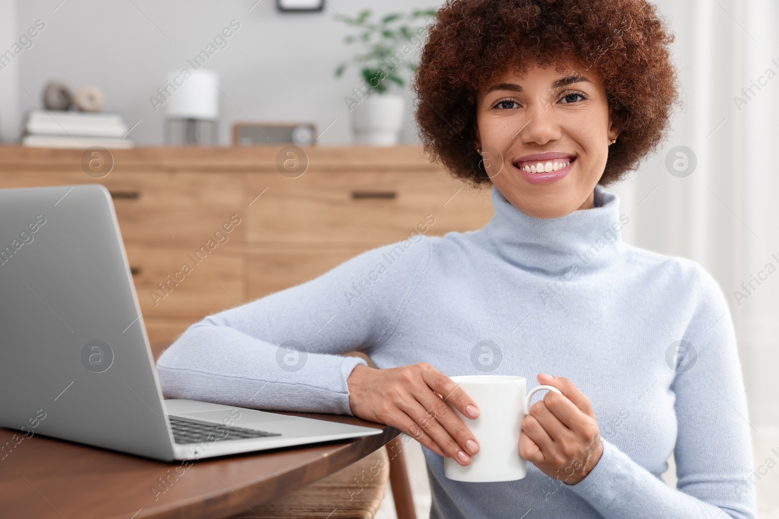 Photo of Beautiful young woman using laptop and drinking coffee at wooden table in room