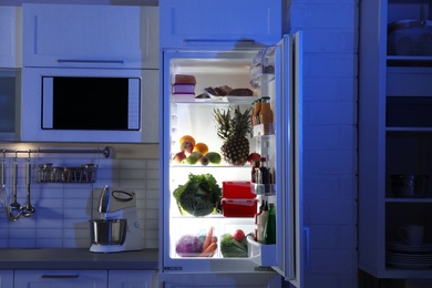 Photo of Open refrigerator full of products in stylish kitchen interior at night