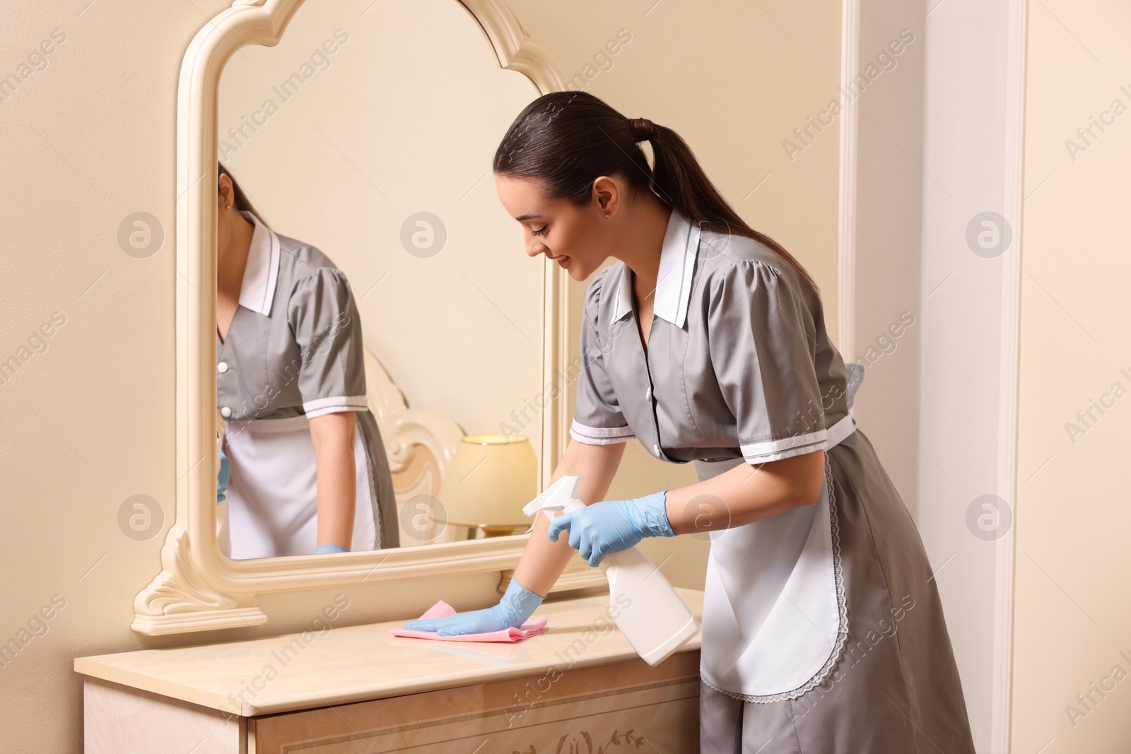 Photo of Young chambermaid cleaning dressing table in hotel room