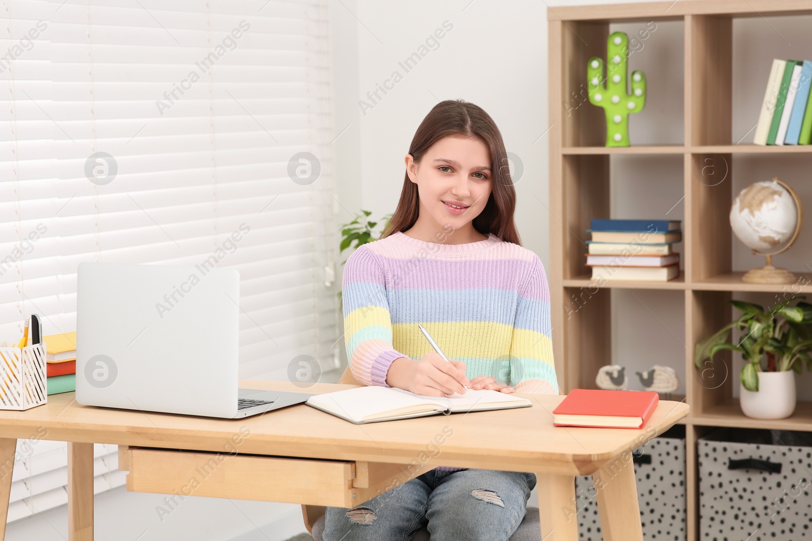 Photo of Cute girl writing in notepad near laptop at desk in room. Home workplace