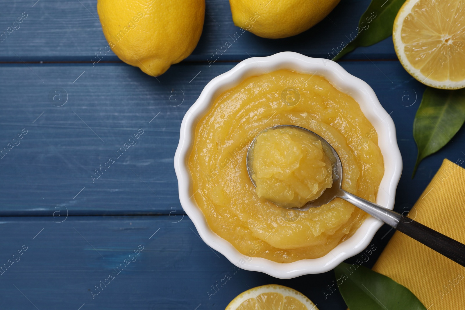 Photo of Delicious lemon curd in bowl, fresh citrus fruits and spoon on blue wooden table, flat lay. Space for text