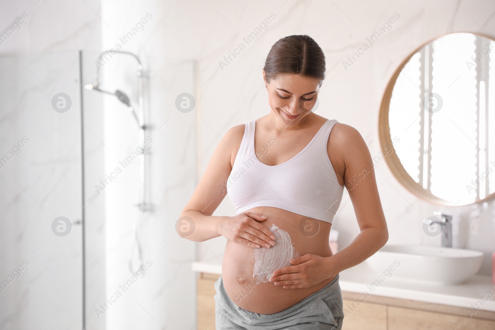 Photo of Young pregnant woman applying cosmetic product on belly in bathroom