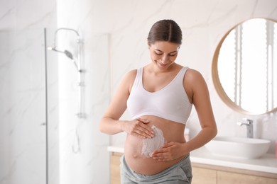 Photo of Young pregnant woman applying cosmetic product on belly in bathroom