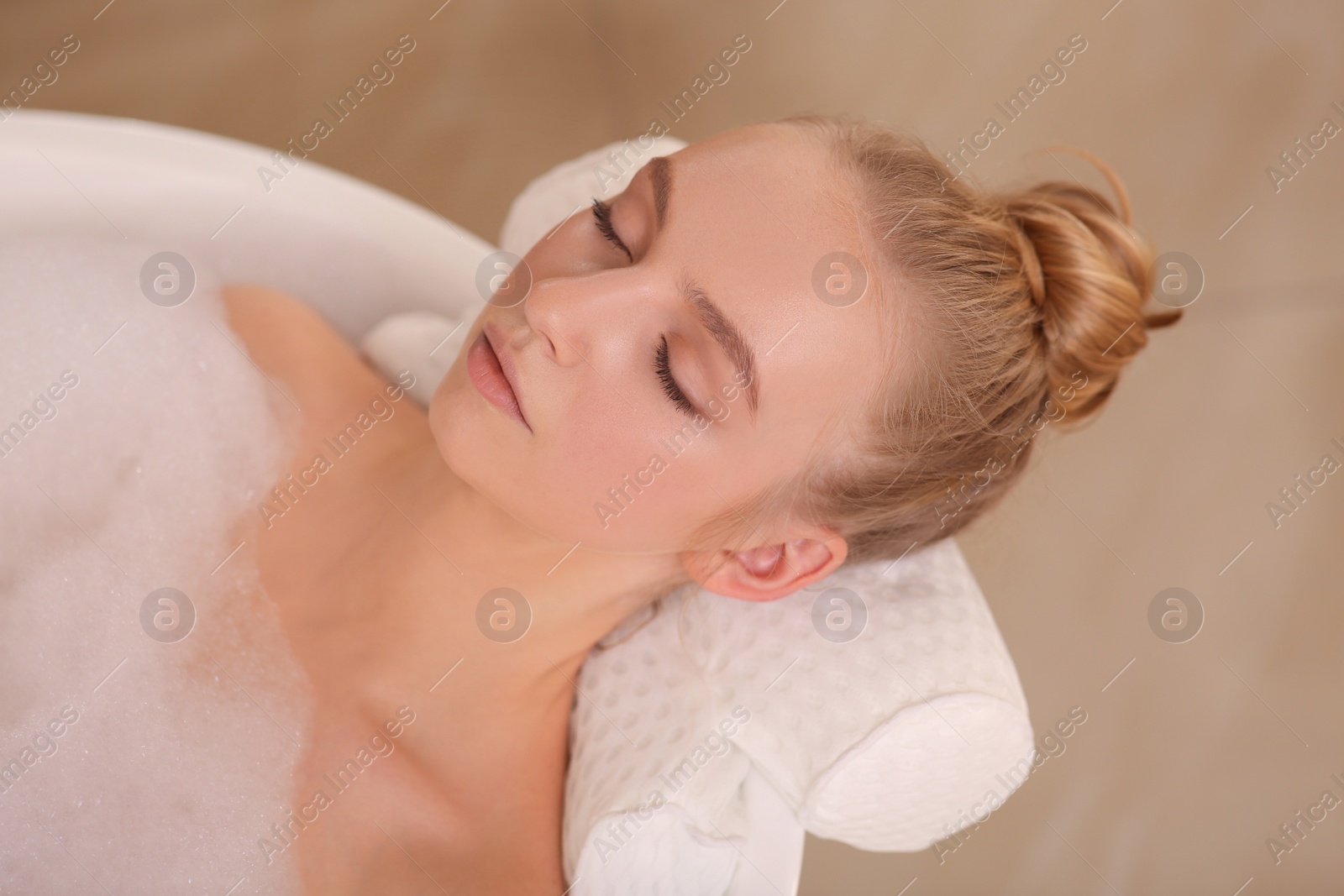 Photo of Young woman using pillow while enjoying bubble bath indoors