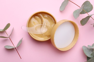 Photo of Under eye patches in jar with spatula and green twigs on light pink background, flat lay. Cosmetic product