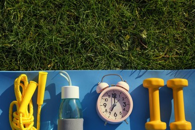 Photo of Alarm clock, skipping rope, dumbbells, bottle of water and fitness mat on green grass, flat lay with space for text. Morning exercise