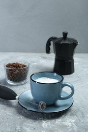 Photo of Mini mixer (milk frother), whipped milk in cup and coffee beans on grey textured table