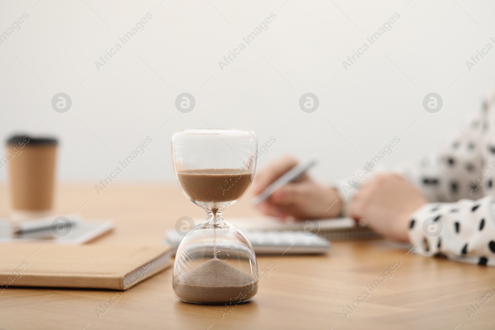 Photo of Hourglass with flowing sand on desk. Woman taking notes while using calculator indoors, selective focus