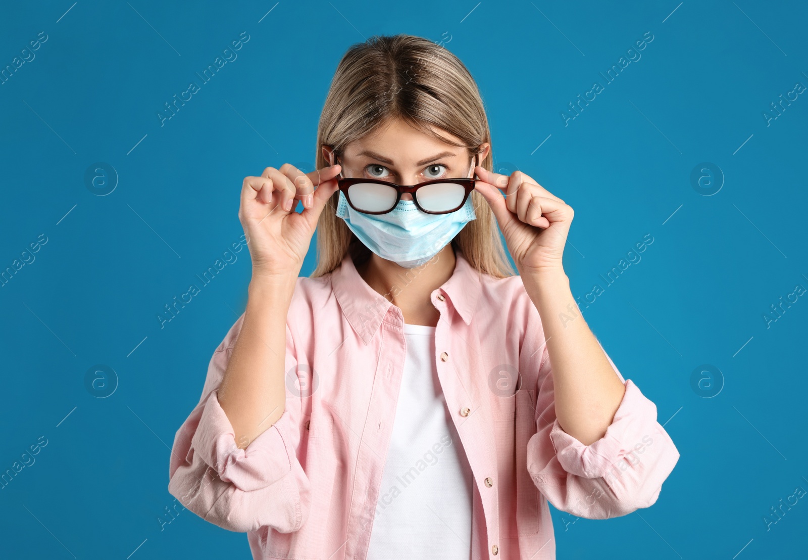 Photo of Woman with foggy glasses caused by wearing disposable mask on blue background. Protective measure during coronavirus pandemic