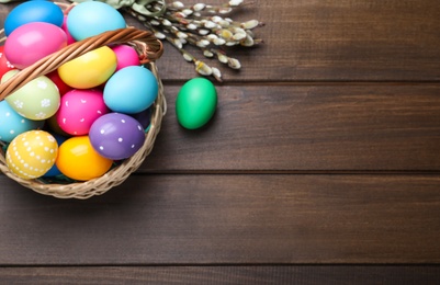 Photo of Colorful Easter eggs in wicker basket and willow branches on wooden table, flat lay. Space for text