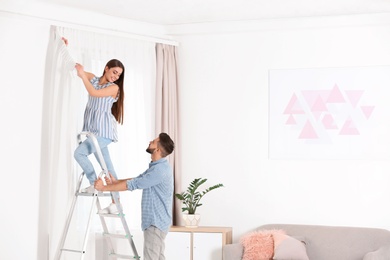 Young couple hanging window curtain in room. Space for text