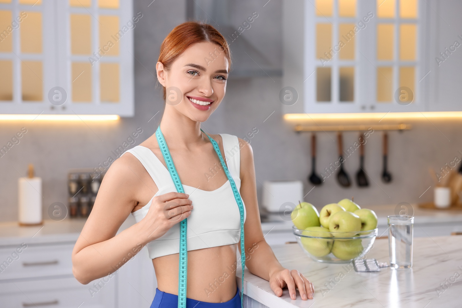 Photo of Slim woman with measuring tape near table with pills, glass of water and apples in kitchen. Weight loss