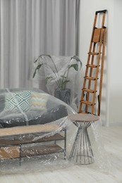 Photo of Modern sofa, table, houseplant covered with plastic film and step ladder at home