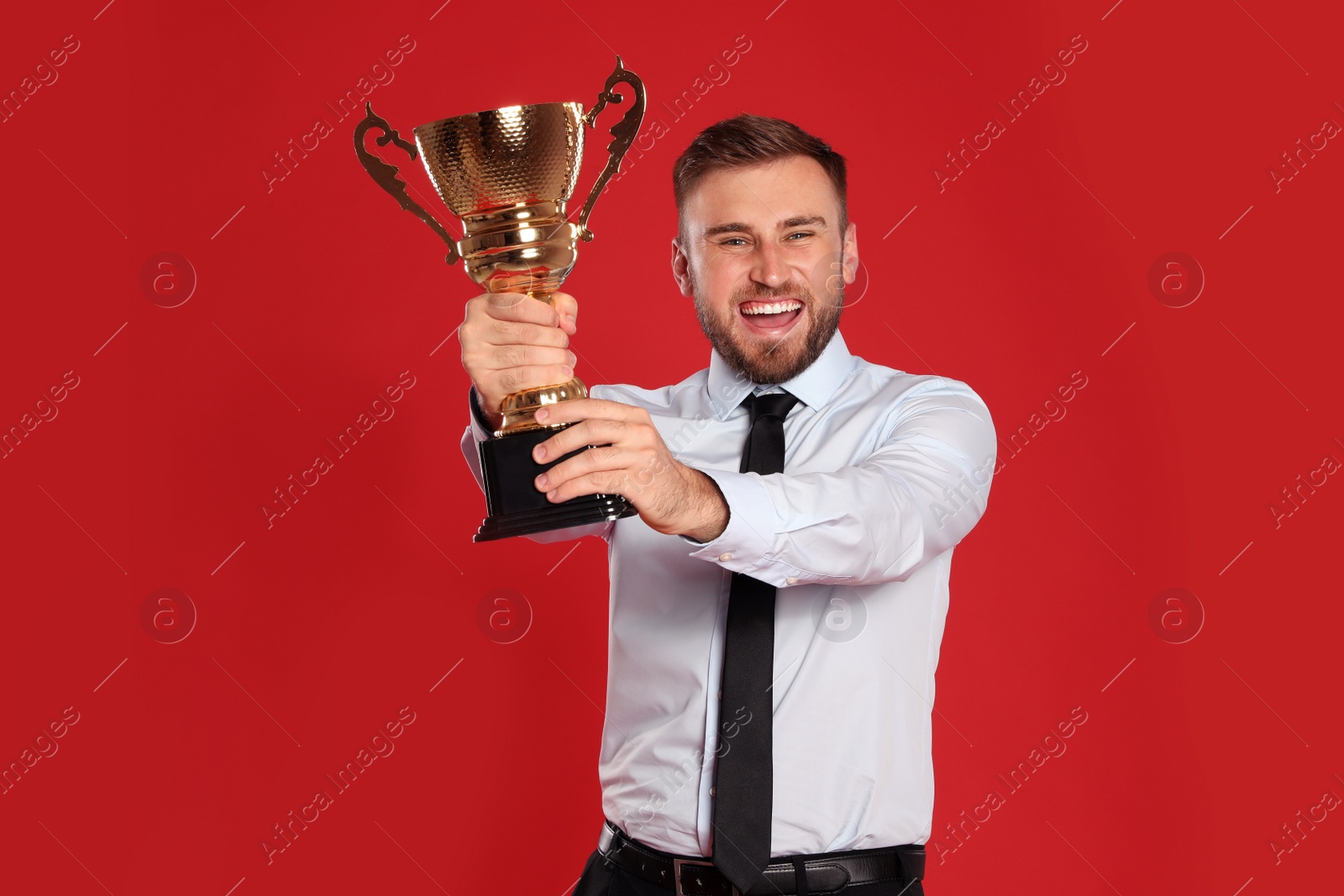 Photo of Portrait of happy young businessman with gold trophy cup on red background