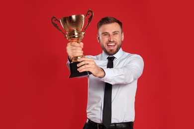 Photo of Portrait of happy young businessman with gold trophy cup on red background