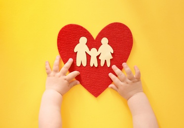 Photo of Little child's hands near red heart and paper silhouette of people on color background, top view