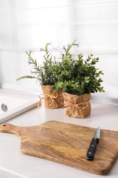 Photo of Artificial potted herbs, board and knife on white marble countertop in kitchen. Home decor