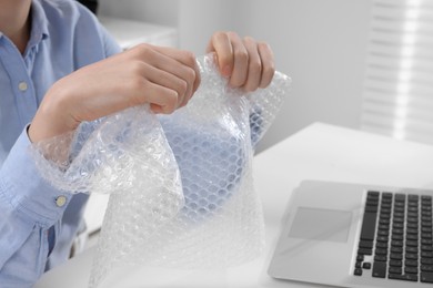 Woman popping bubble wrap at table in office, closeup. Stress relief