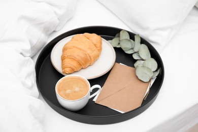 Tray with tasty croissant, cup of coffee and envelope on white bed