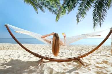 Image of Woman with refreshing cocktail relaxing in hammock under green palm leaves on beach