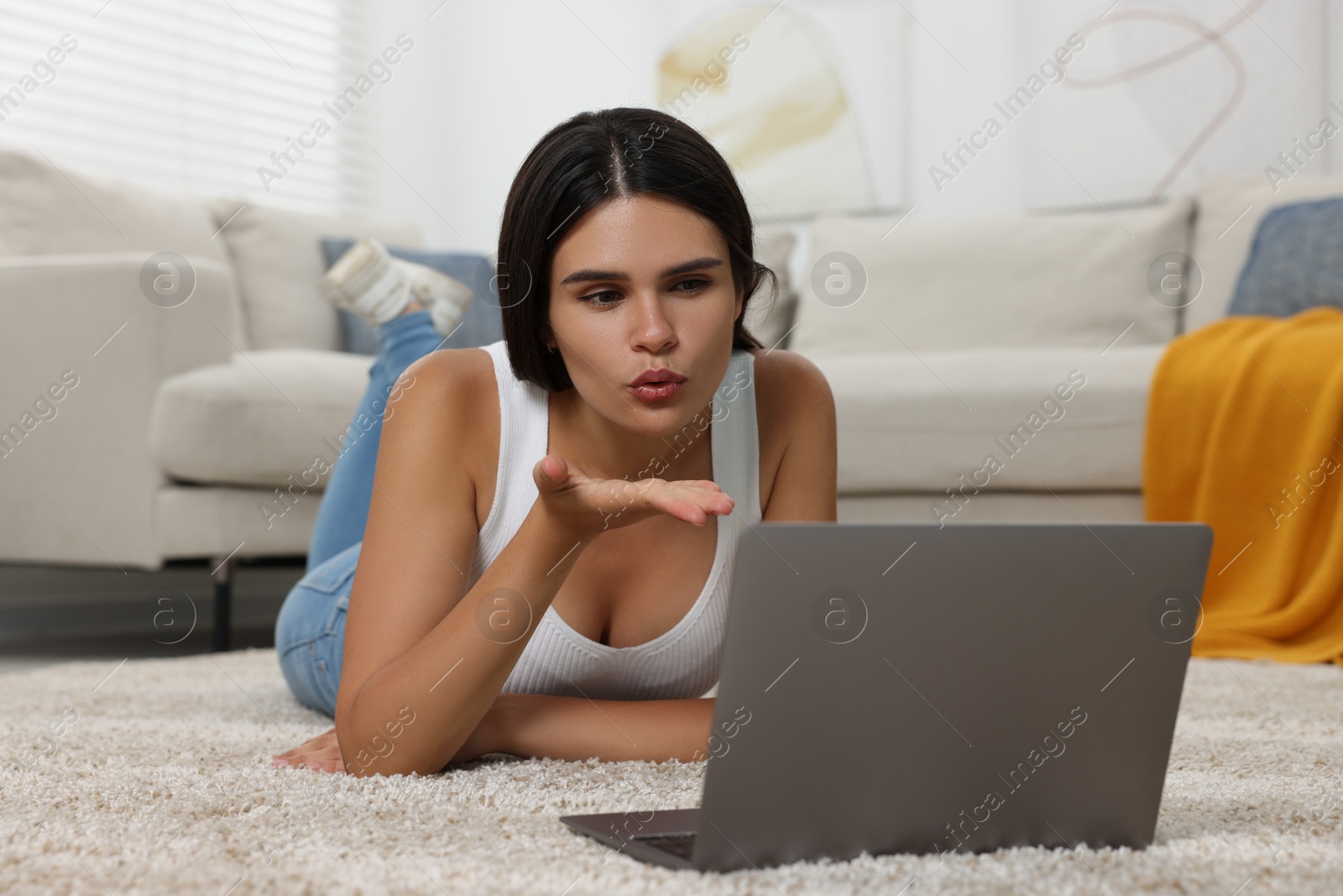 Photo of Young woman having video chat via laptop and blowing kiss on floor at home