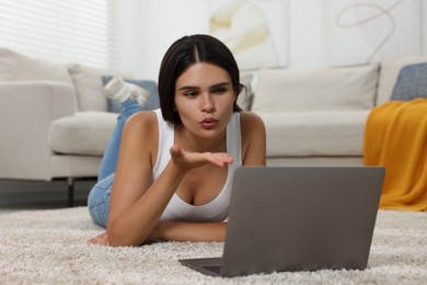 Photo of Young woman having video chat via laptop and blowing kiss on floor at home