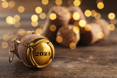 Image of Cork of sparkling wine and muselet cap with engraving 2023 on wooden table, space for text. Bokeh effect