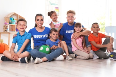 Photo of Happy volunteers with children sitting on floor indoors