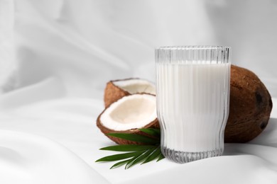 Glass of delicious coconut milk, palm leaves and coconuts on white fabric, space for text