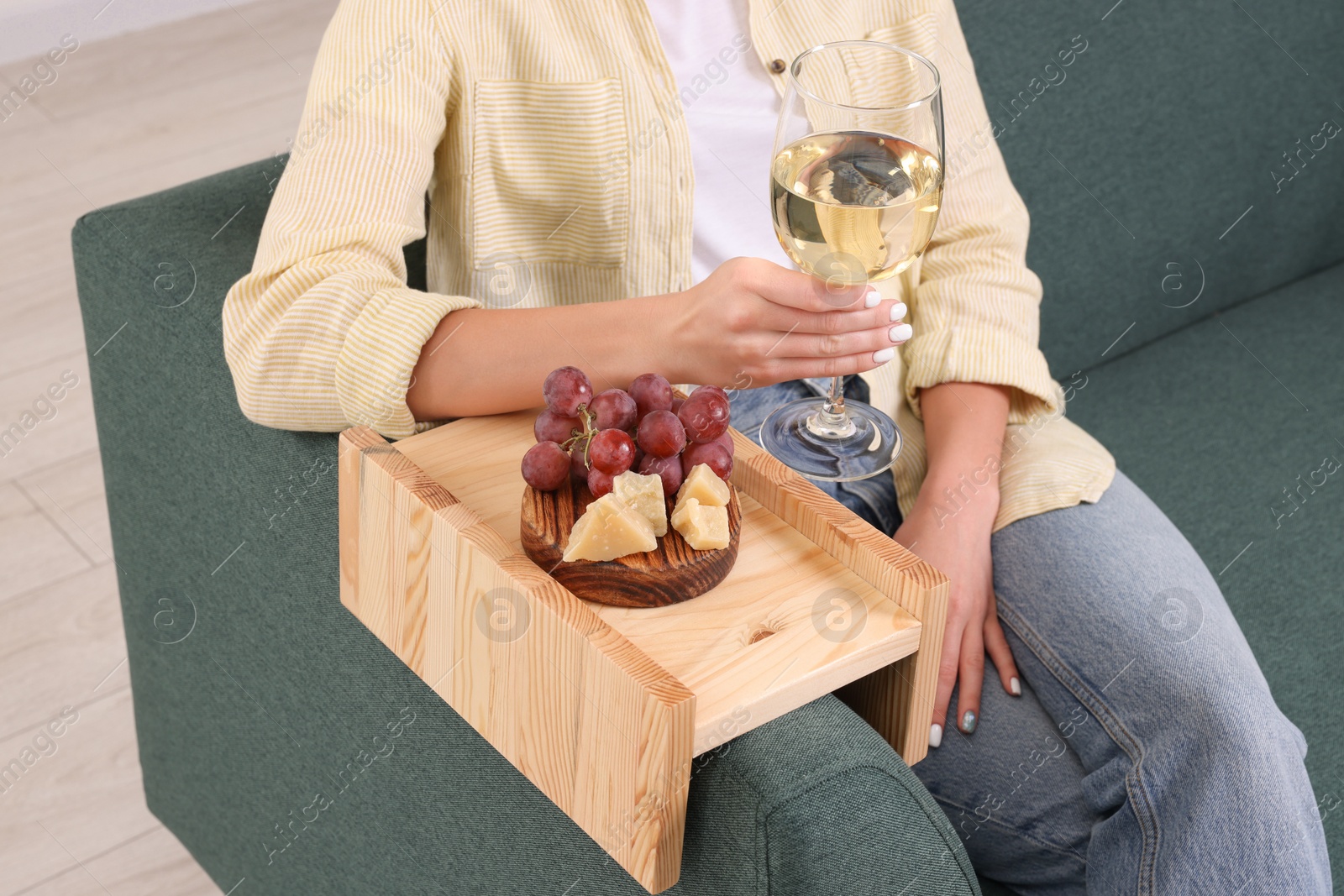 Photo of Grapes and cheese on sofa armrest wooden table. Woman holding glass of wine at home, closeup