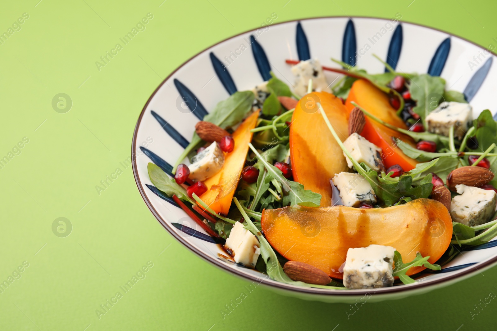 Photo of Tasty salad with persimmon, blue cheese, pomegranate and almonds served on light green background, closeup. Space for text