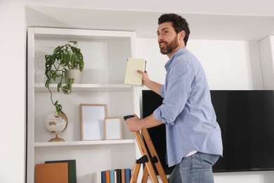 Photo of Man on wooden folding ladder taking books from shelf at home