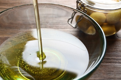 Pouring fresh olive oil into bowl on wooden background, closeup