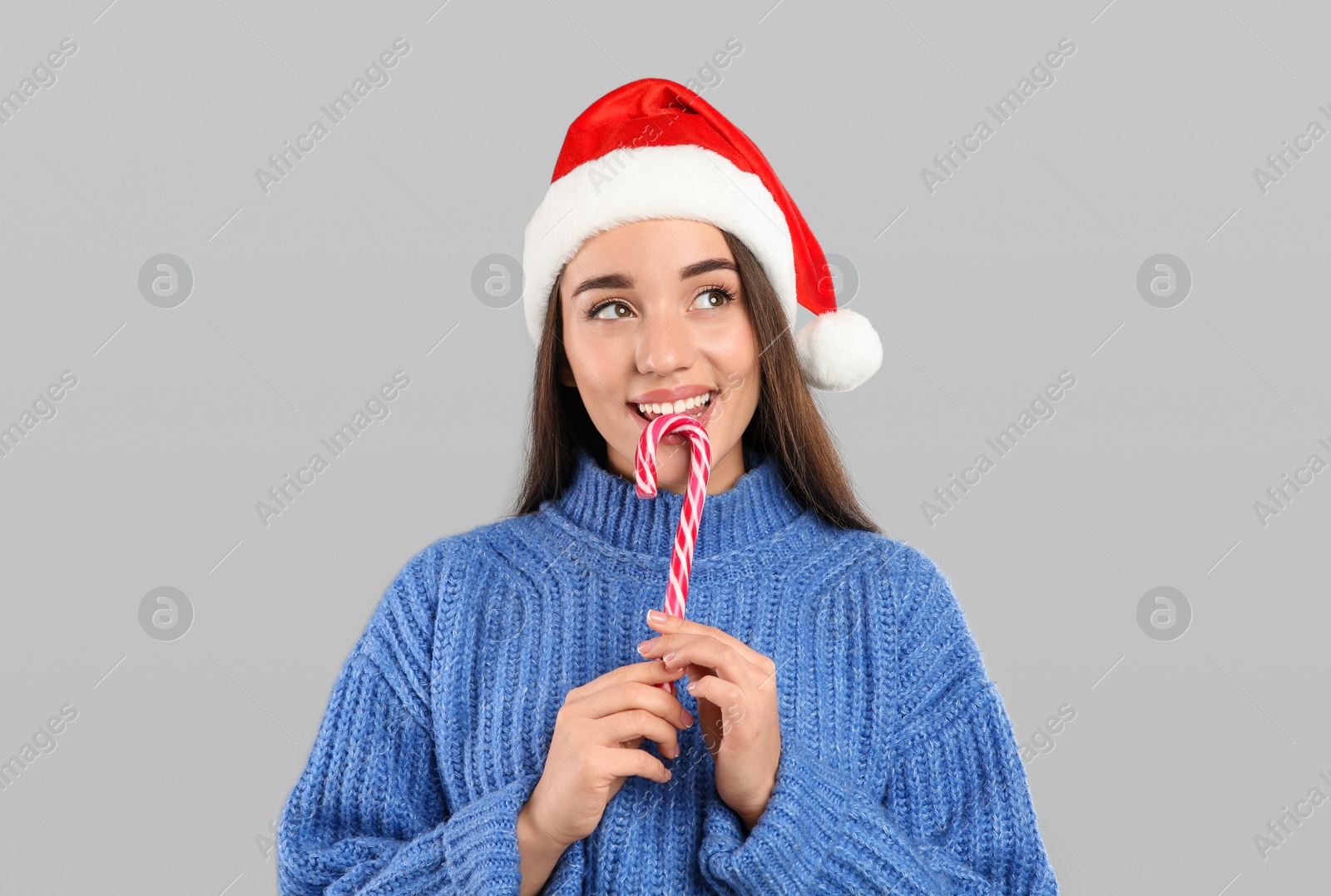 Photo of Young woman in blue sweater and Santa hat holding candy cane on grey background. Celebrating Christmas