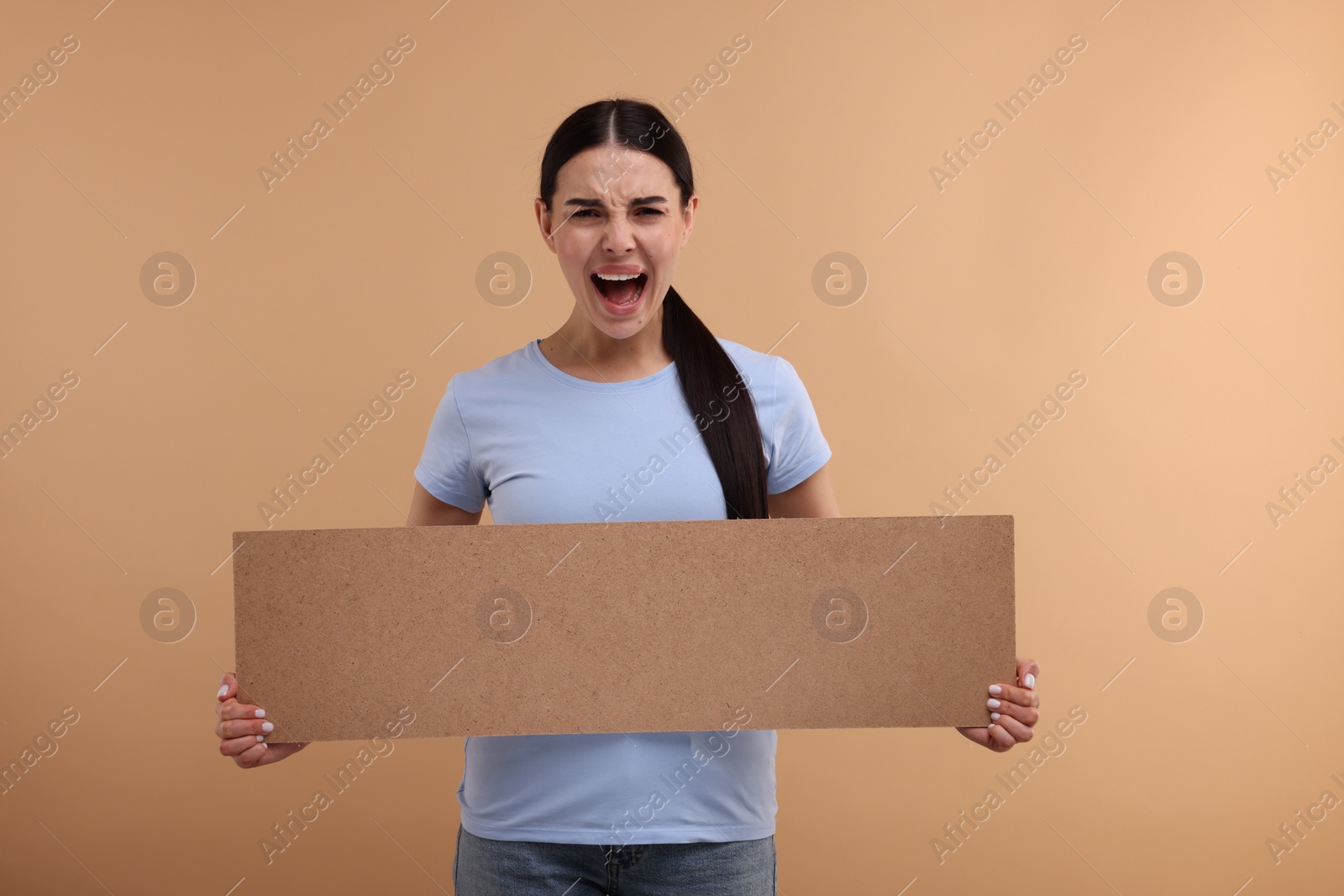 Photo of Angry woman holding blank cardboard banner on beige background, space for text