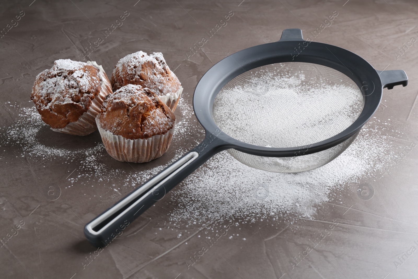 Photo of Sieve with sugar powder and muffins on grey textured table, closeup