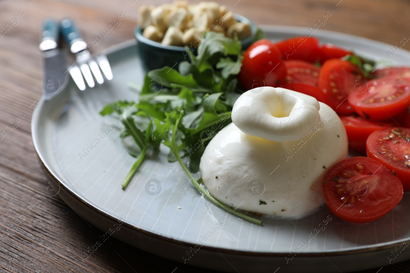 Photo of Delicious burrata cheese with tomatoes and arugula served on wooden table, closeup