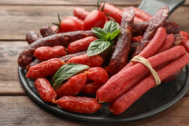 Photo of Different thin dry smoked sausages, basil and tomatoes on wooden table, closeup