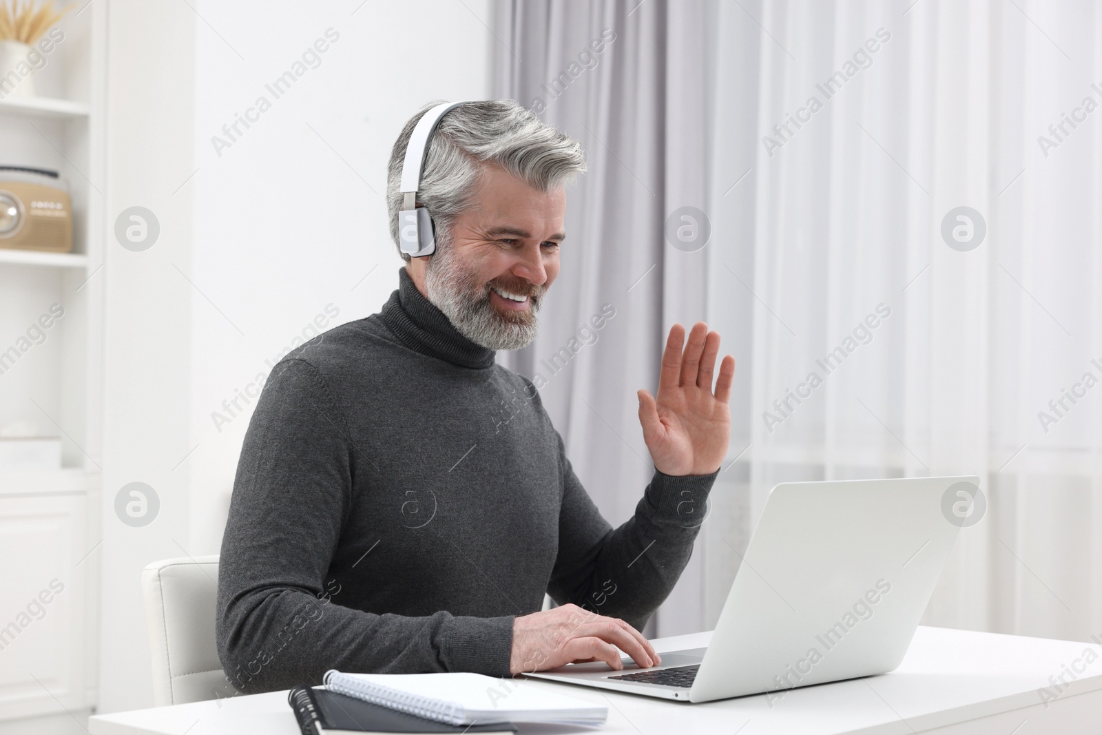 Photo of Man in headphones waving hello during video chat via laptop at home