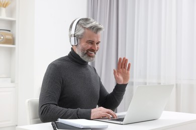 Photo of Man in headphones waving hello during video chat via laptop at home