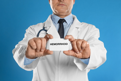 Doctor holding business card with word HEMORRHOID on blue background, closeup