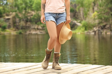 Young woman with hat on wooden pier near lake, focus on legs. Camping season