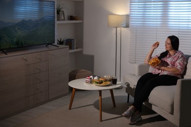 Photo of Happy overweight woman with chips watching TV on sofa at home