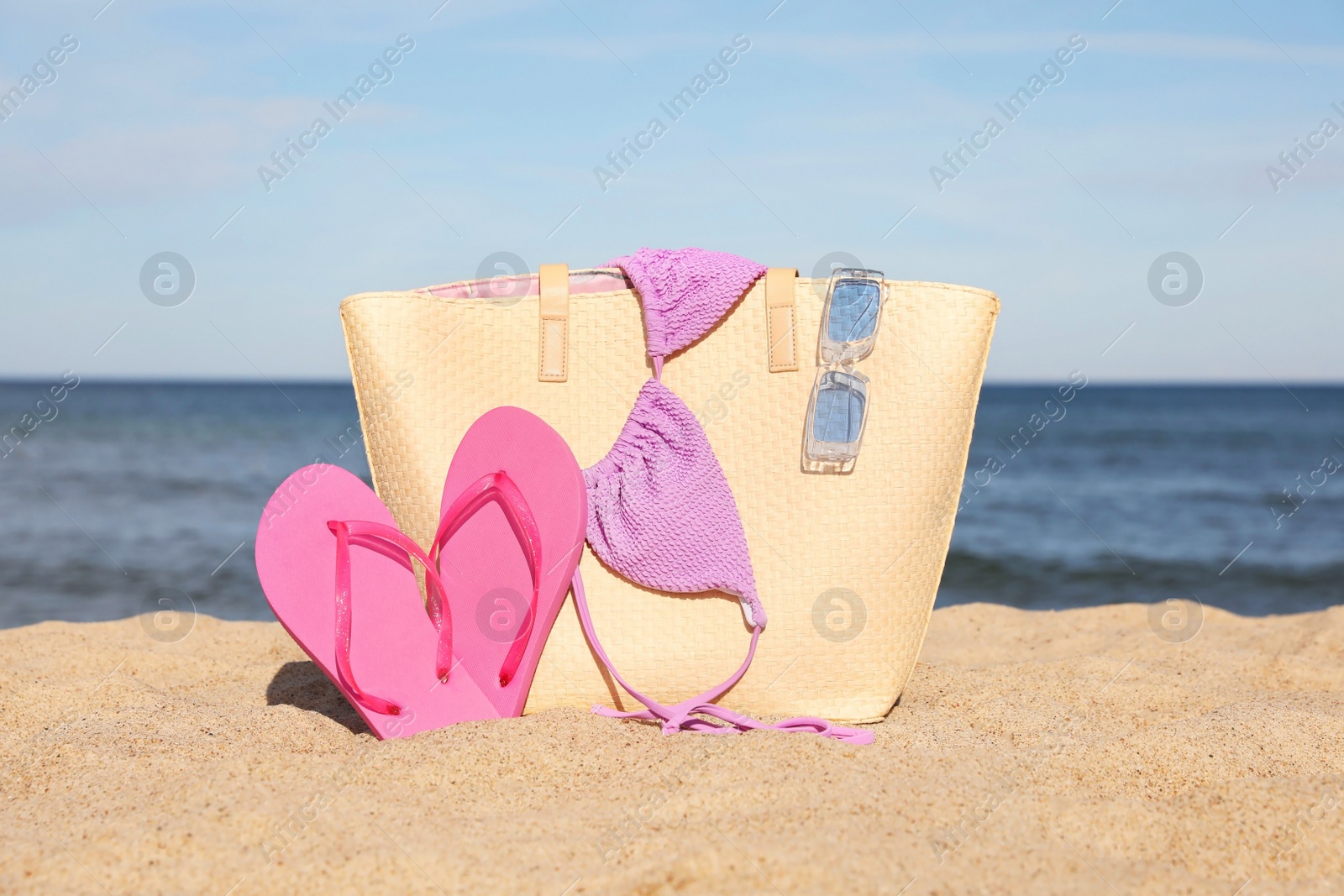 Photo of Summer bag with slippers, bikini top and sunglasses on sand near sea